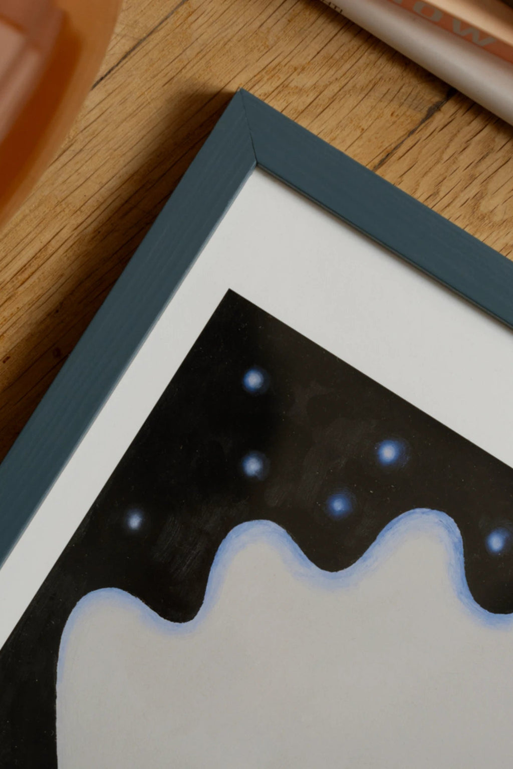 The Poster Club Sternenkind, an abstract art print featuring a dark black background with a light grey wavy organic shape and glowing blue celestial dots, framed in a matte dark blue frame with a clean white mat. This modern art print is perfect as whimsical wall art for a contemporary bedroom, nursery, or gallery wall.