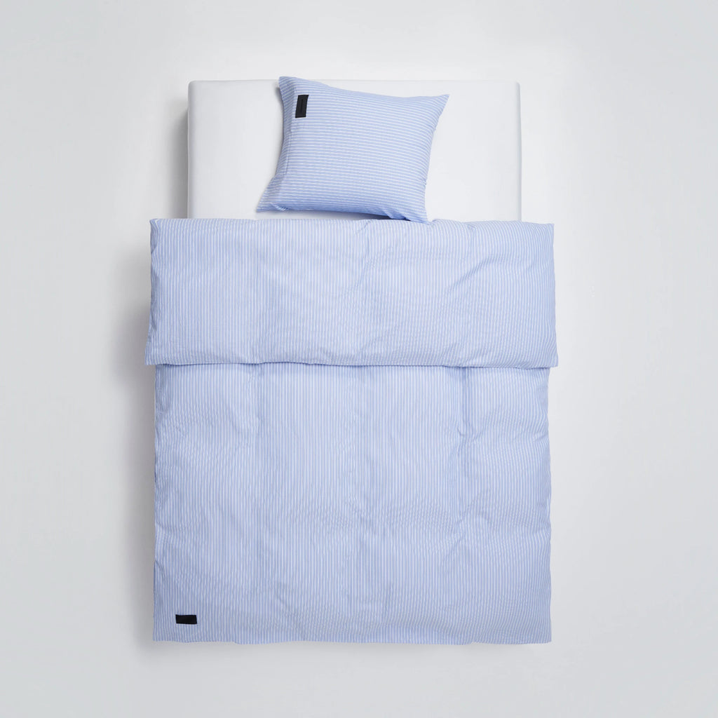Magniberg Wall Street Pillow Case - Stripe Light Blue Oxford, a classic and crisp Oxford cotton pillowcase showcasing fine vertical white stripes against a soft light blue background, complete with a subtle black brand label. This durable, stylish bed linen offers a fresh, modern minimalist aesthetic for Scandinavian-inspired bedrooms or contemporary guest rooms seeking a timeless look.