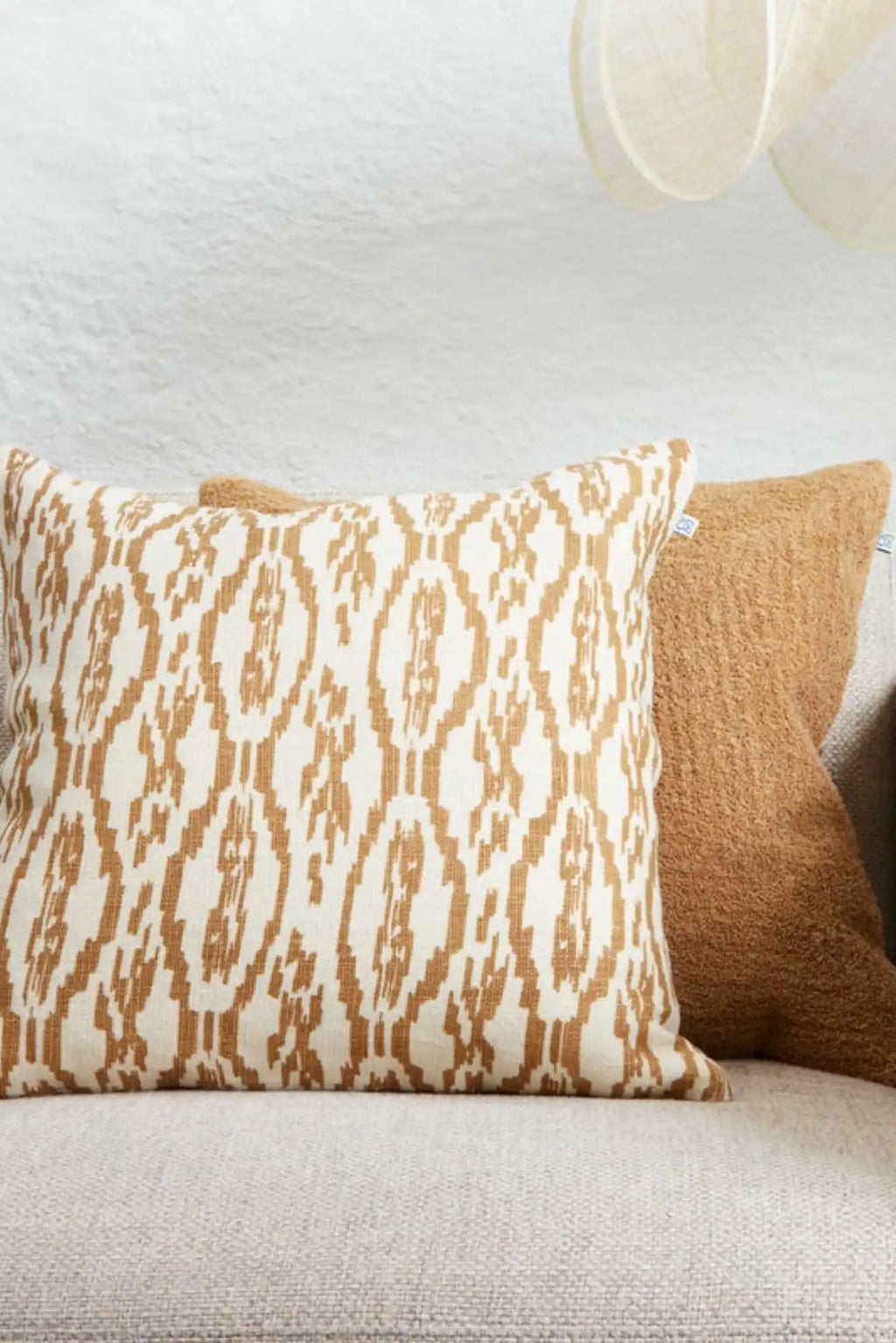 Chhatwal & Jonsson Deccan Linen Throw Pillow, a square woven linen cushion featuring an intricate cream and warm brown ikat pattern with a natural, slightly slubby texture. This bohemian-inspired accent pillow is perfect for adding global chic style to a living room sofa or bedroom decor, often paired with a plush caramel brown textured cushion.