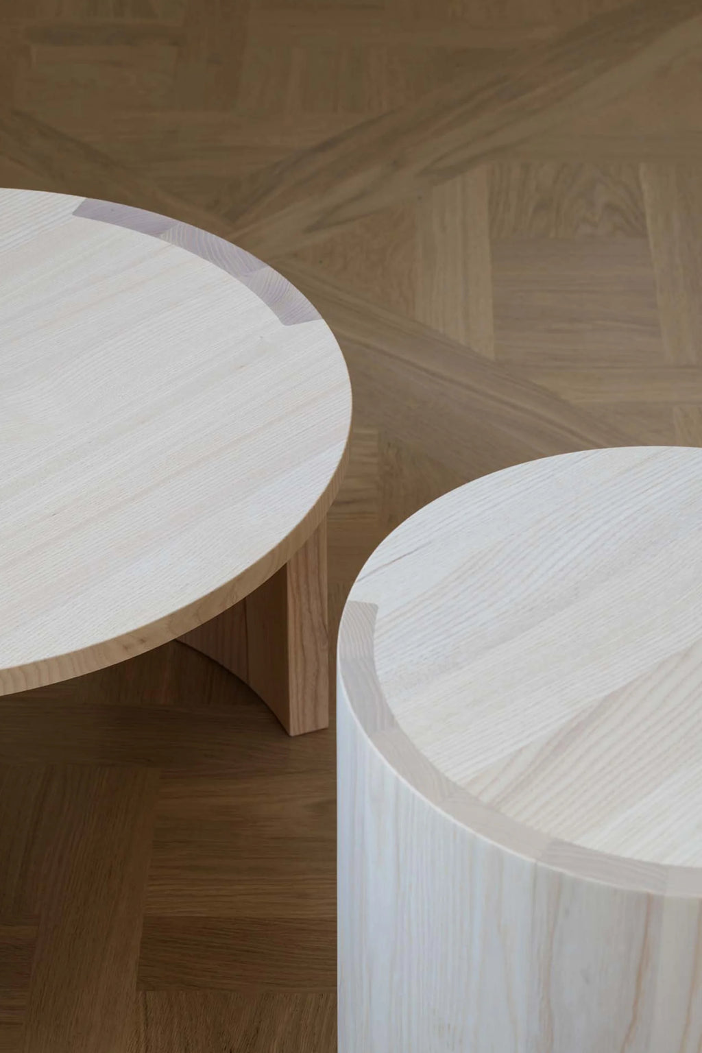Made by Choice Airisto Side Table, a set of two modern minimalist round side tables crafted from pale solid ash wood with a visible natural grain, featuring one cylindrical form and one with a curved, intersecting base. These light wood tables are perfect as Scandinavian living room furniture or contemporary bedroom accent tables.