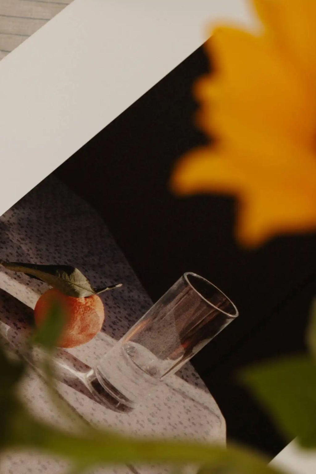 The Poster Club Sunrise, a warm-toned still life poster featuring a vibrant, blurred orange and yellow abstract element set against a deep dark background. This photographic art print showcases a clear cylindrical glass and a single orange fruit with a green leaf, resting on a subtly textured light surface.