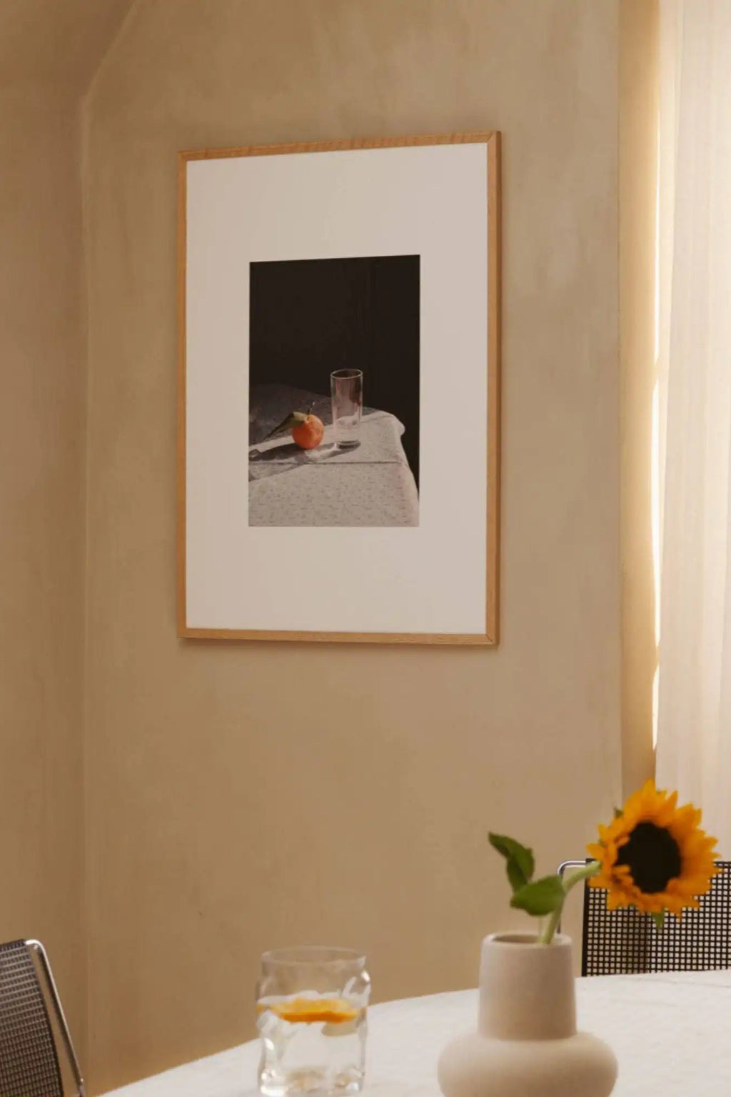 The Poster Club Sunrise, a contemporary still life art print featuring a warm-toned peach or tangerine and a clear glass on a textured white tablecloth against a dark, moody background, framed in light natural oak with a crisp white mat. Ideal for modern minimalist dining rooms, organic modern kitchens, or contemporary living spaces.