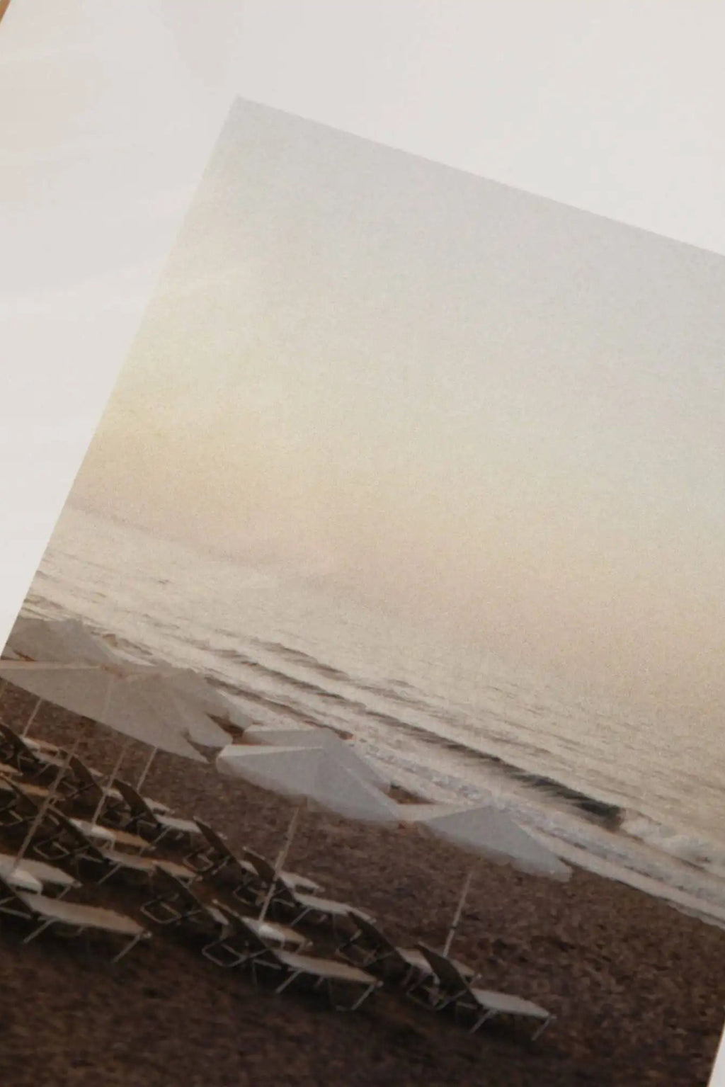 The Poster Club Sunset Swims, a serene fine art print capturing a tranquil beach landscape at dusk. The minimalist photography features neat rows of white beach umbrellas and dark lounge chairs on sandy shores, with soft ocean waves meeting a muted, warm-toned sky. This calming coastal wall art is ideal for creating a peaceful ambiance in Scandinavian, organic modern, or contemporary bedrooms and living spaces.