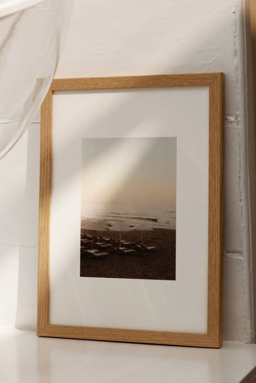 The Poster Club Sunset Swims, a serene coastal photography print capturing a warm sunset over a calm sea, featuring white beach umbrellas and lounge chairs on a sandy shore in soft peachy and brown tones. This minimalist art print is presented with a crisp white mat and framed in a light natural oak frame. Ideal as tranquil wall art for a coastal living room, serene bedroom, or contemporary gallery wall.
