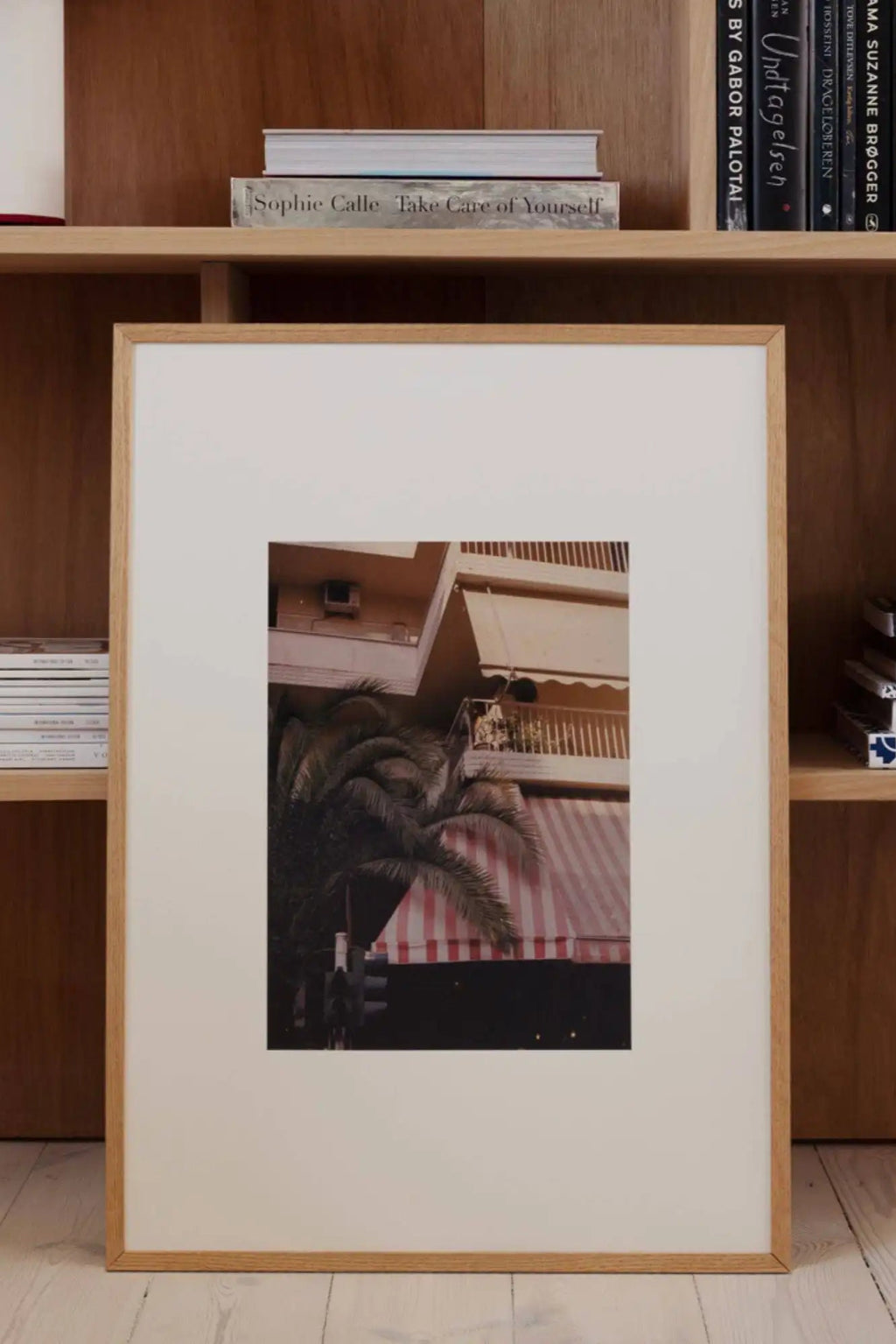 The Poster Club The Corner We Both Loved, a vintage-style photographic art print featuring a warm, faded Mediterranean street scene with terracotta buildings, a cream balcony awning, and a red and white striped awning peeking through dark palm fronds. This framed poster print is presented with a crisp white mat and a light natural oak frame, offering a clean, contemporary aesthetic. Perfect for creating a summery gallery wall or adding a touch of nostalgic travel art to a modern living room or bedroom.