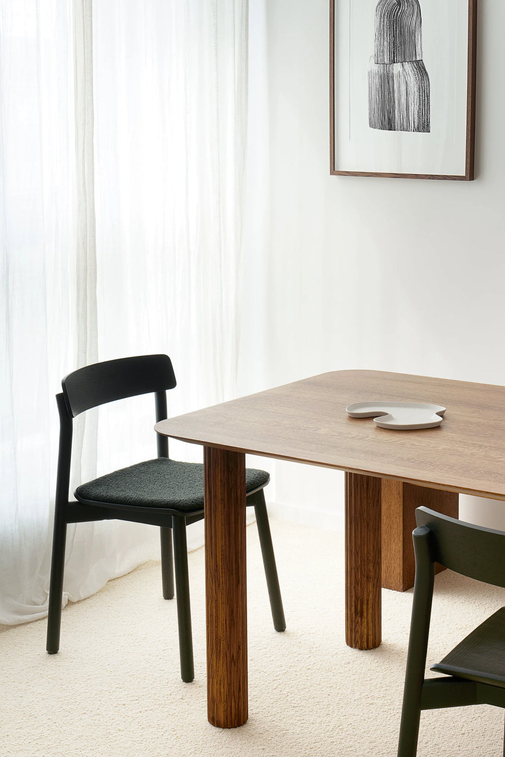 TAMO Filar Dining Table featuring a smooth, light brown oak wood top with rounded corners and fluted, cylindrical legs. A gray, organic-shaped plate rests on the table, surrounded by sleek, dark wood dining chairs with textured black fabric seats, all on a cream-colored carpet. This premium table is ideal for modern dining room settings.