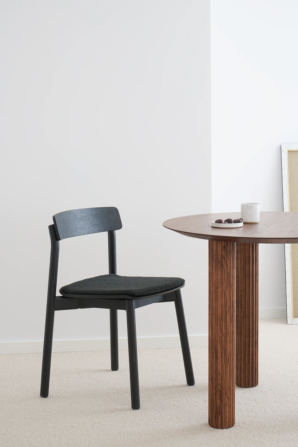TAMO Arche Chair, displaying its sleek matte black oak frame, curved backrest, and textured dark gray upholstered seat pad, next to a modern wood table with snacks in a lifestyle setting. This premium black wooden dining chair is perfect for minimalist interiors.