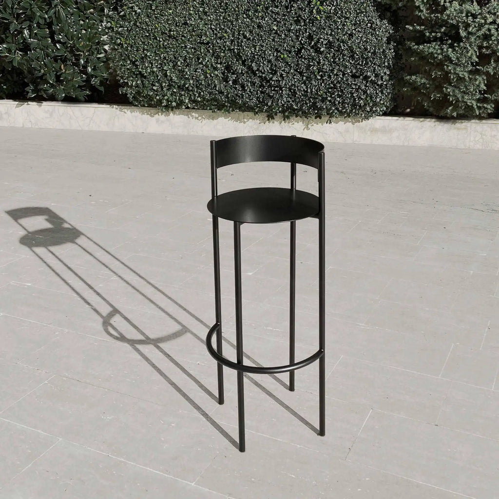 Around Works Pub Stool, a sleek matte black metal bar stool with a modern minimalist design, featuring a circular seat, a low curved backrest, and slender cylindrical legs with a round footrest. This contemporary bar height stool is perfect for kitchen islands, home bars, or industrial chic spaces.