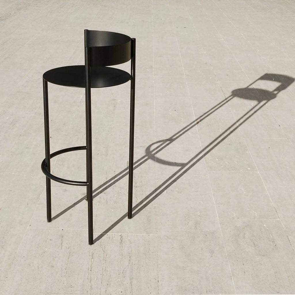 Around Works Pub Stool, a modern minimalist matte black metal bar stool featuring a sleek, slender cylindrical frame, a comfortable round seat, and a low curved backrest, complete with a circular footrest. This contemporary bar stool is perfect for industrial chic kitchen islands or modern home bar setups.
