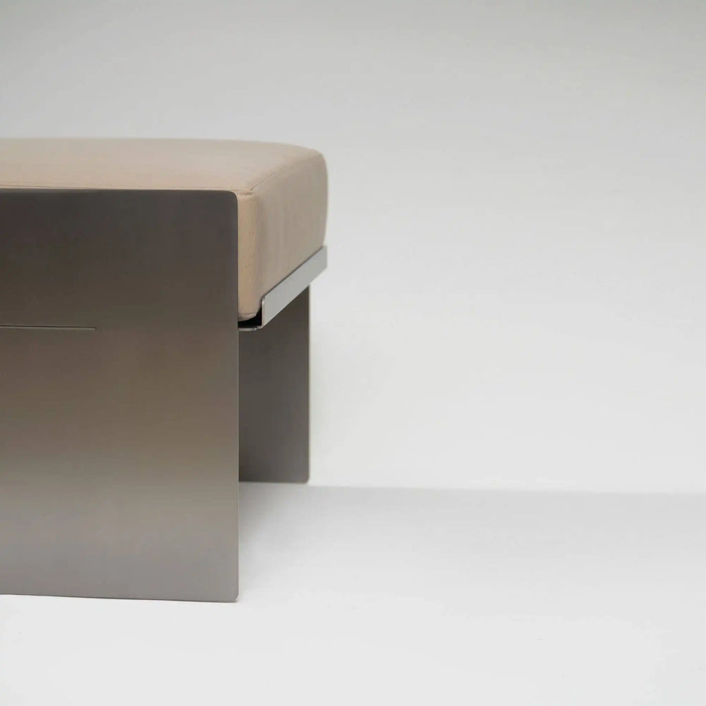 Around Works C.01 Lounge, a modern minimalist pouf featuring a rectangular beige upholstered cushion perched on a sleek matte taupe-grey metal base with a subtle linear detail and a slim silver connecting bracket. This contemporary lounge ottoman is perfect for adding refined seating or a stylish accent to living rooms, bedrooms, or entryways.