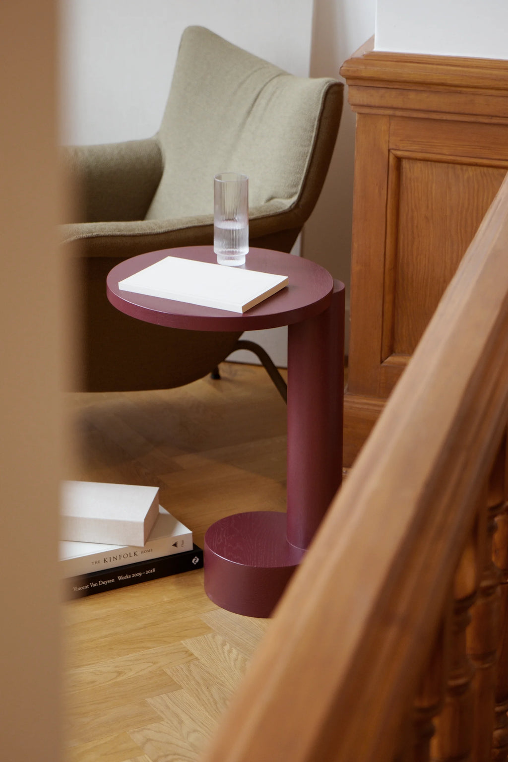 TAMO Bold Side Table, with its rich matte plum/burgundy finish and contemporary c-shape design, features a minimalist round top and base. Adorned with a clear ribbed glass and a white book, this premium accent piece complements a light green armchair in a stylish living room setup. Ideal for modern interior design.