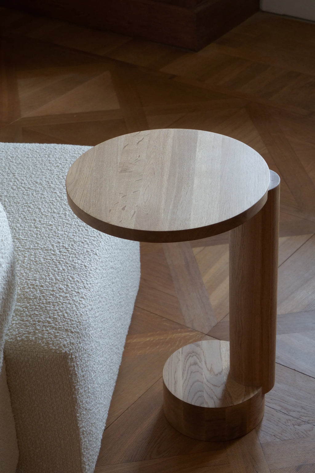 TAMO Bold Side Table, a premium light brown natural oak wood side table with a circular top, and a unique curved base, next to a textured cream sofa on a parquet floor. Ideal for modern living spaces.