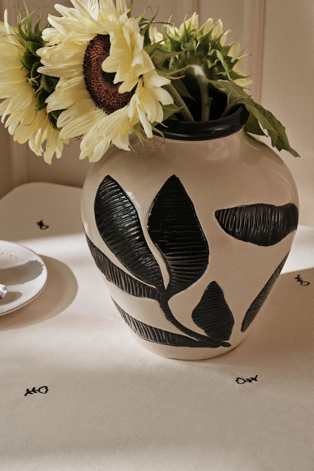 Broste Copenhagen Herbert Vase, a charming cream vase with textured black leaf designs, filled with bright yellow sunflowers, placed on a patterned tablecloth.