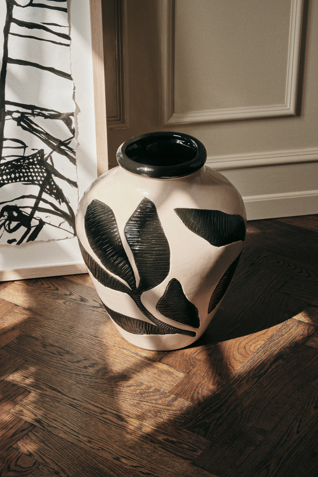 Broste Copenhagen Herbert Vase, a beautiful cream ceramic vase with raised black leaf patterns, sitting on a warm wooden floor, bathed in natural sunlight.