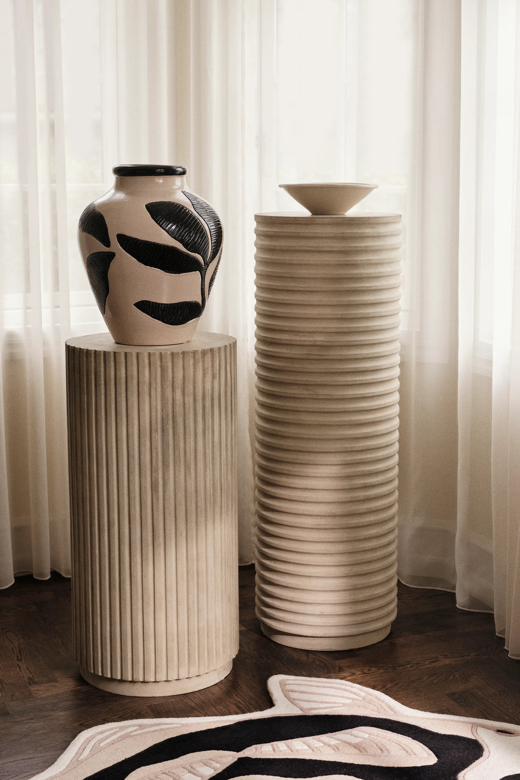 Broste Copenhagen Herbert Vase, a striking cream vase with textured black leaf patterns, displayed on a fluted pedestal next to sheer white curtains in a sunlit room.