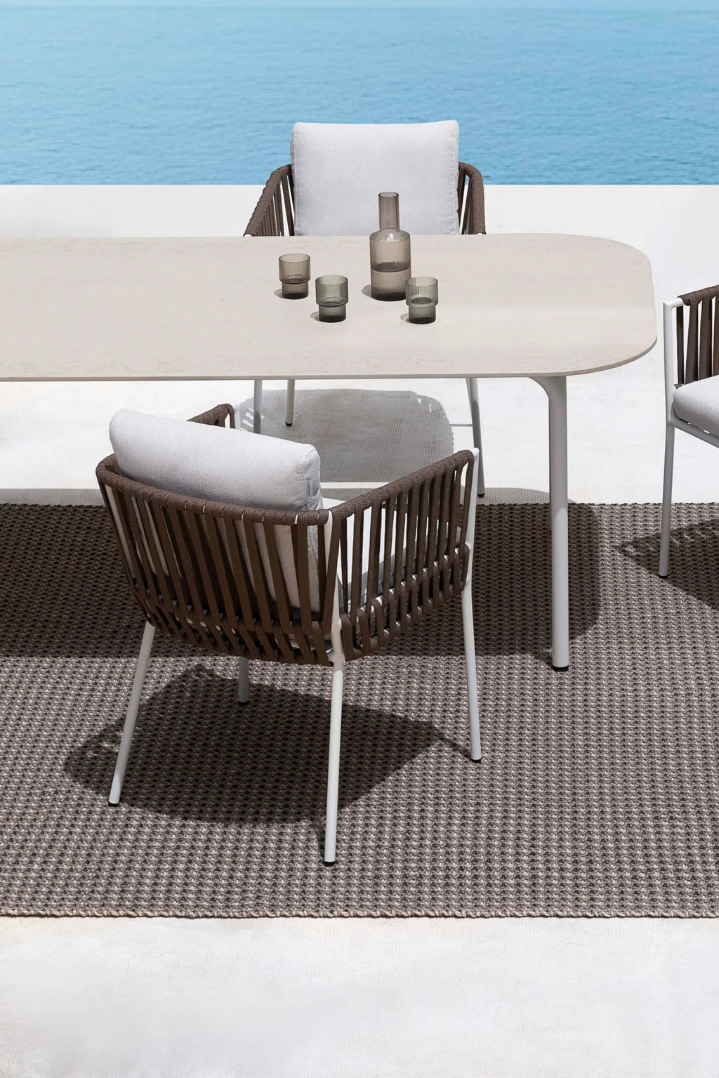 Gandia Blasco Corda Outdoor Dining Chair, a modern outdoor chair featuring a sturdy white metal frame with a curved backrest and armrests crafted from intricate brown woven rope. This contemporary design includes plush light grey outdoor fabric cushions for ultimate comfort and a refined aesthetic.