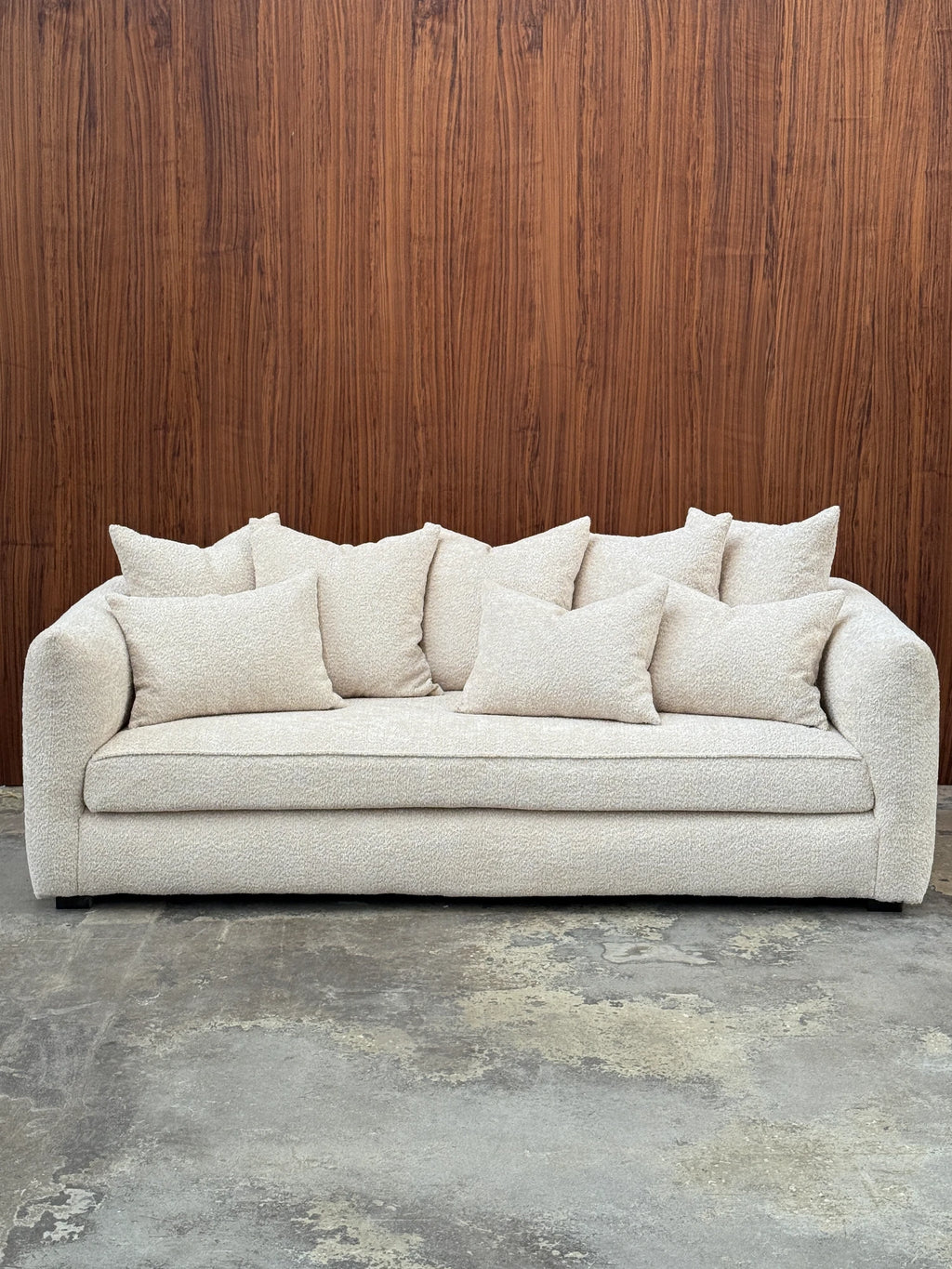 Tinted Objects Cornell Sofa, a contemporary organic modern sofa upholstered in luxurious cream bouclé fabric with a plush, textured weave and inviting deep seating. Its gently curved arms and generous proportions are complemented by eight matching throw pillows, creating a cozy and sophisticated silhouette. Ideal for minimalist living rooms, modern apartments, or creating a warm, inviting lounge area.