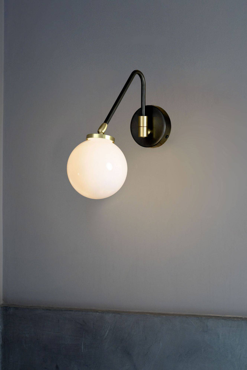 CTO Lighting Array Single Wall Lamp, a sleek modern wall light featuring a matte black metal curved arm and a round backplate, accented with elegant brushed brass detailing. It showcases a luminous spherical opal glass globe diffuser, providing soft ambient light for contemporary interiors. This minimalist wall sconce is perfect for bedroom accent lighting, a chic hallway fixture, or a stylish living room statement piece in industrial chic or mid-century modern spaces.