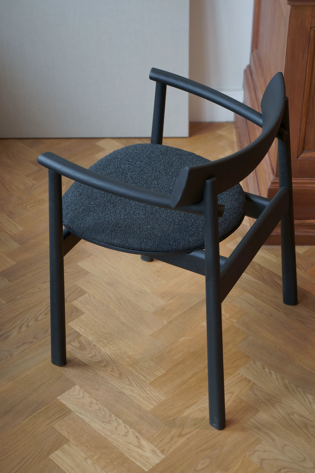 TAMO Elipsa Chair, a modern matte black wooden armchair with sleek curved arms and a textured dark grey upholstered round seat, displayed on a herringbone hardwood floor. Ideal for contemporary living spaces.