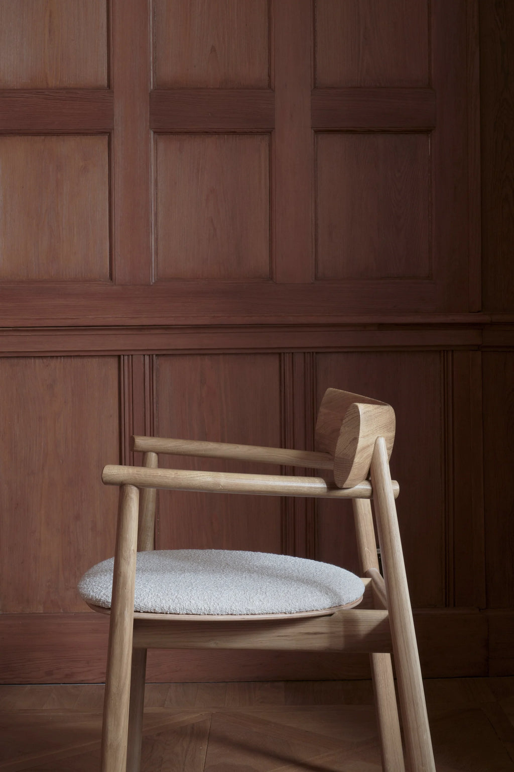 TAMO Elipsa Chair, a premium light oak wood chair with a natural grain finish and a round, light grey textured boucle fabric upholstered seat, displayed against rich brown paneling. Ideal for modern dining or accent seating.