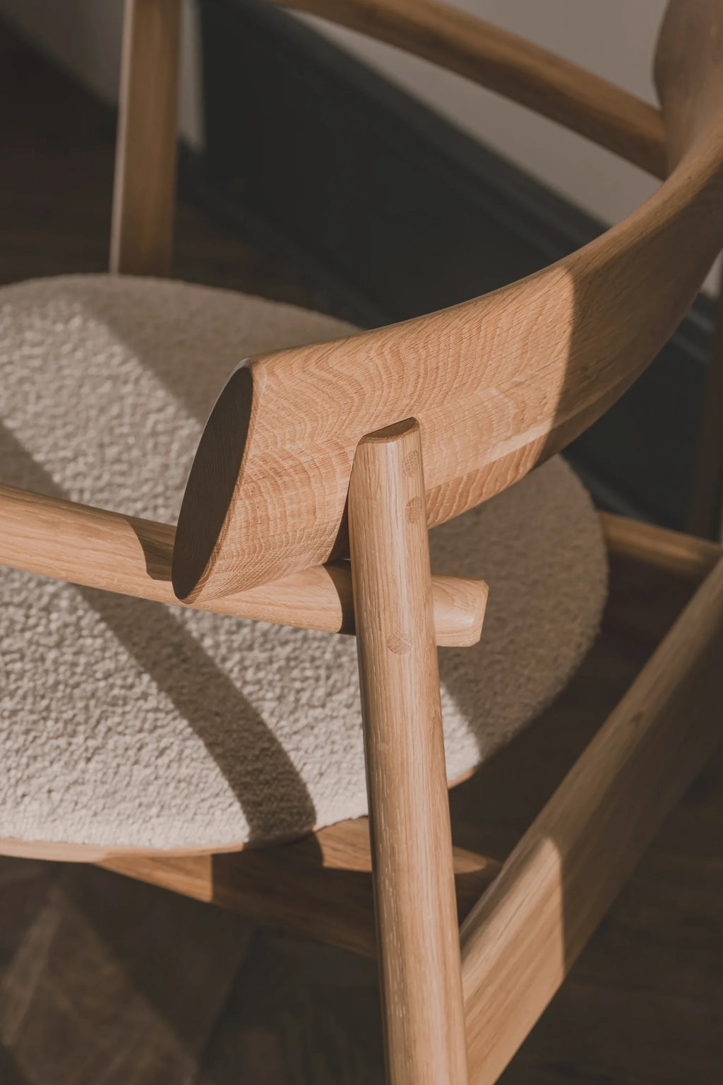 TAMO Elipsa Chair, showcasing its light oak wood frame with visible grain details and a round, textured creme bouclé seat in a sunlit close-up. A luxury modern chair for sophisticated living spaces.