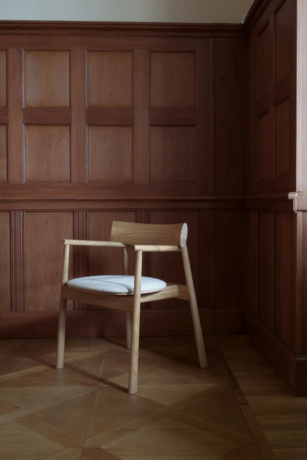 TAMO Elipsa Chair, a premium light oak wood armchair featuring a curved backrest and a light gray, textured upholstered seat, against dark wood-paneled walls. Ideal for luxury dining or office settings.