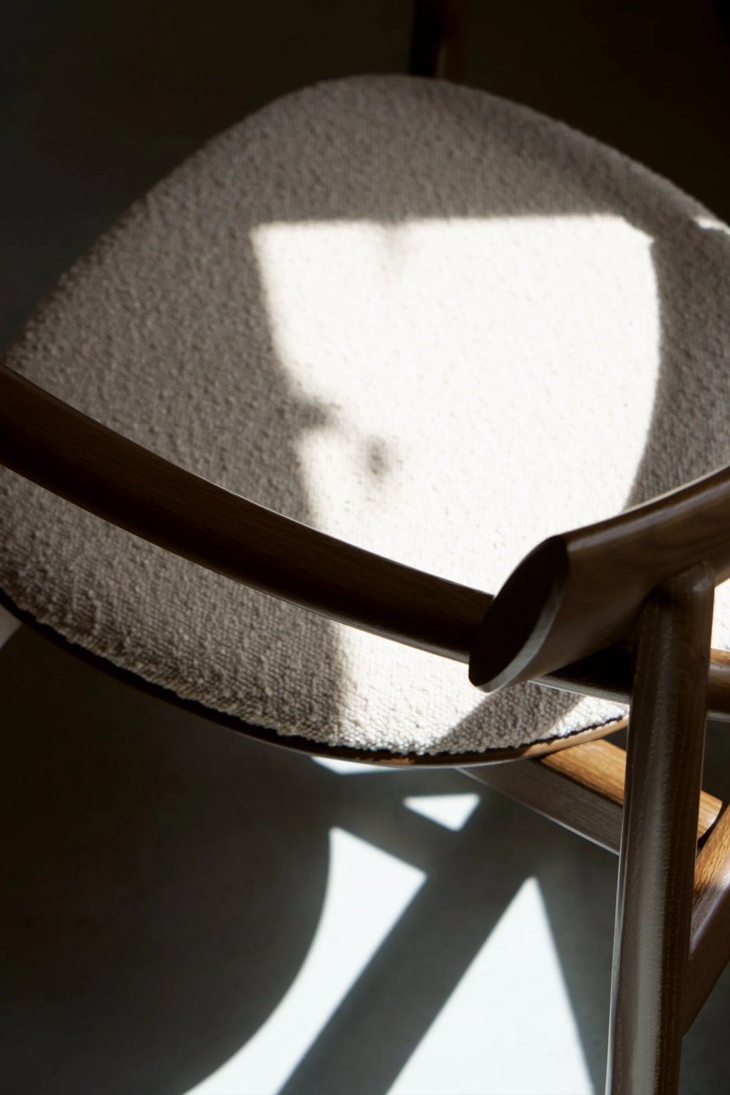 TAMO Elipsa Chair, showcasing a curved, light brown wooden frame and textured, off-white upholstered seat in dappled sunlight. This luxury, handcrafted chair offers sophisticated style. Ideal for modern living spaces.