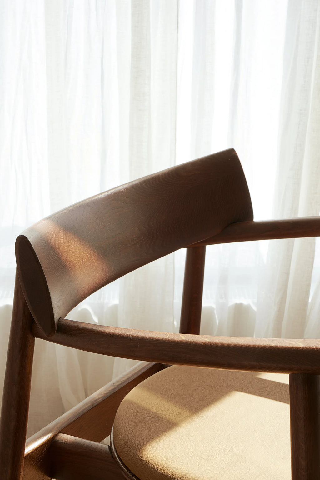 TAMO Elipsa Chair, a close-up of the rich dark brown wood grain frame and curved backrest, with a light tan upholstered seat, against sheer white curtains. This authentic mid-century modern piece is ideal for sophisticated dining or office spaces.
