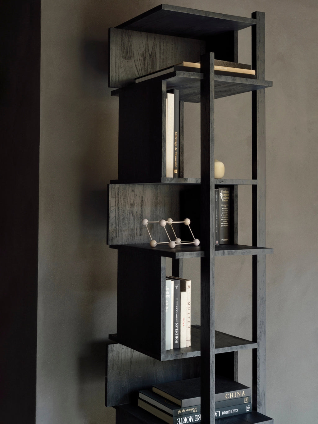 Ethnicraft Abstract Column, a striking dark teak and black bookshelf with a unique, multi-level design, displaying books and a minimalist sculpture, enhancing a contemporary interior.