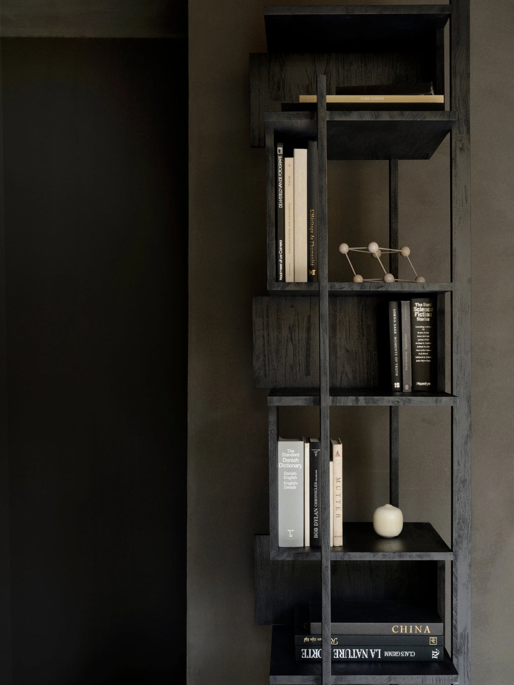 Ethnicraft Abstract Column, a tall, dark teak and black bookshelf with an asymmetrical, geometric design, featuring various books and decorative objects, set against a dark, textured wall.