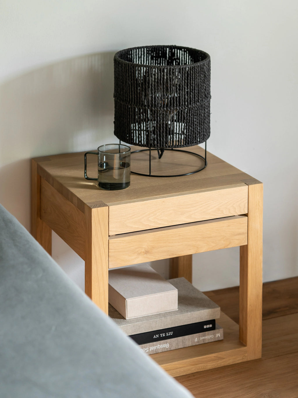 Ethnicraft Azur Bedside Table in light oak, featuring a single drawer and open shelf, styled with a black woven lamp and glass of water, in a modern bedroom setting.