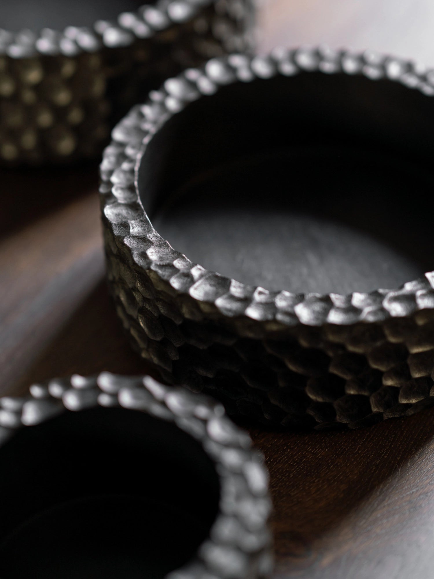 Ethnicraft Chopped Bowl Set, a close-up, high-angle view of three dark, textured bowls with a unique hammered pattern, showcasing their artisanal craftsmanship and deep, rich color on a dark wooden surface.
