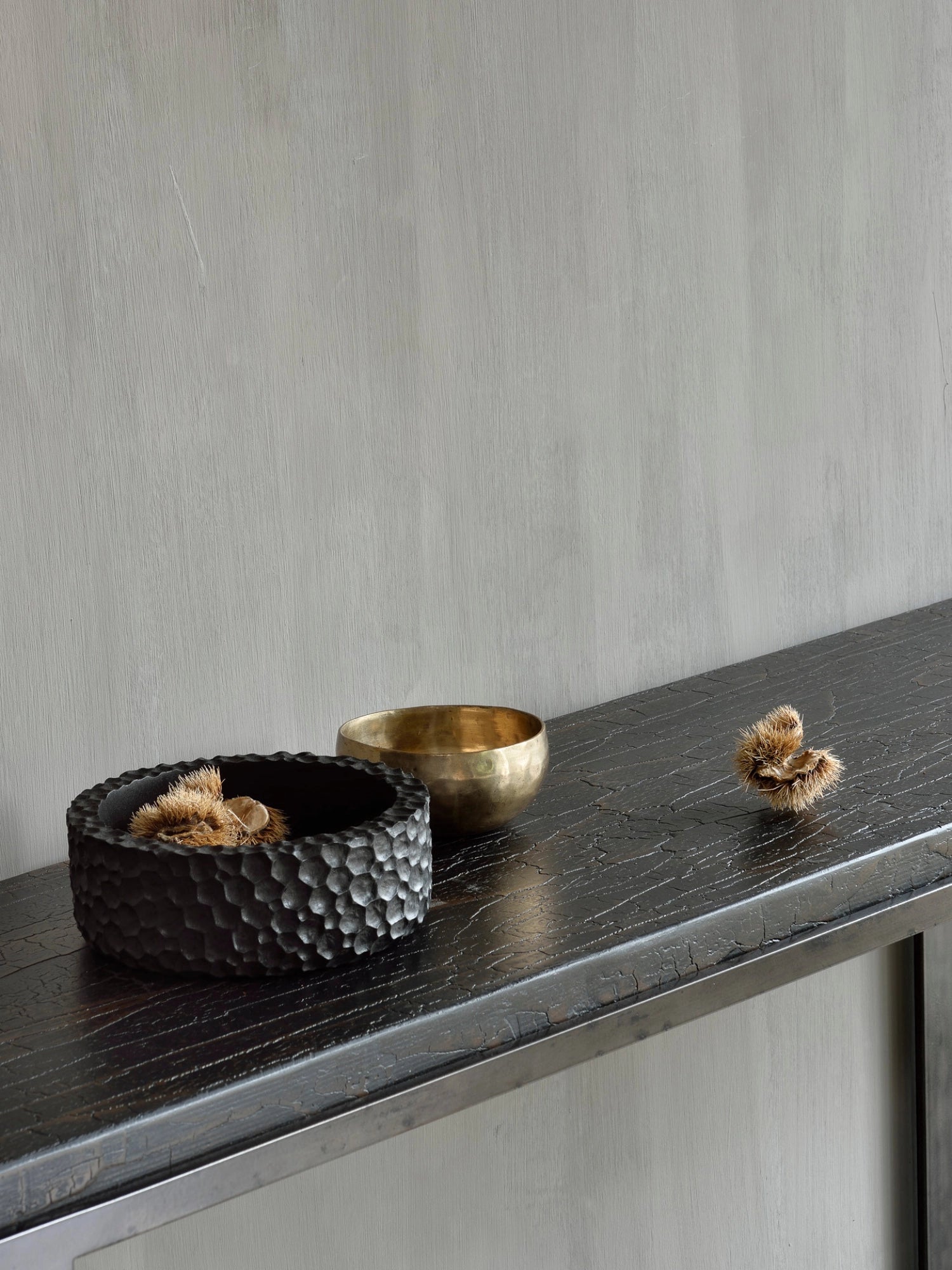Ethnicraft Chopped Bowl Set, featuring a dark, textured bowl filled with spiky seed pods, alongside a smooth brass bowl, all resting on a rustic wooden console table, creating an elegant, natural aesthetic.
