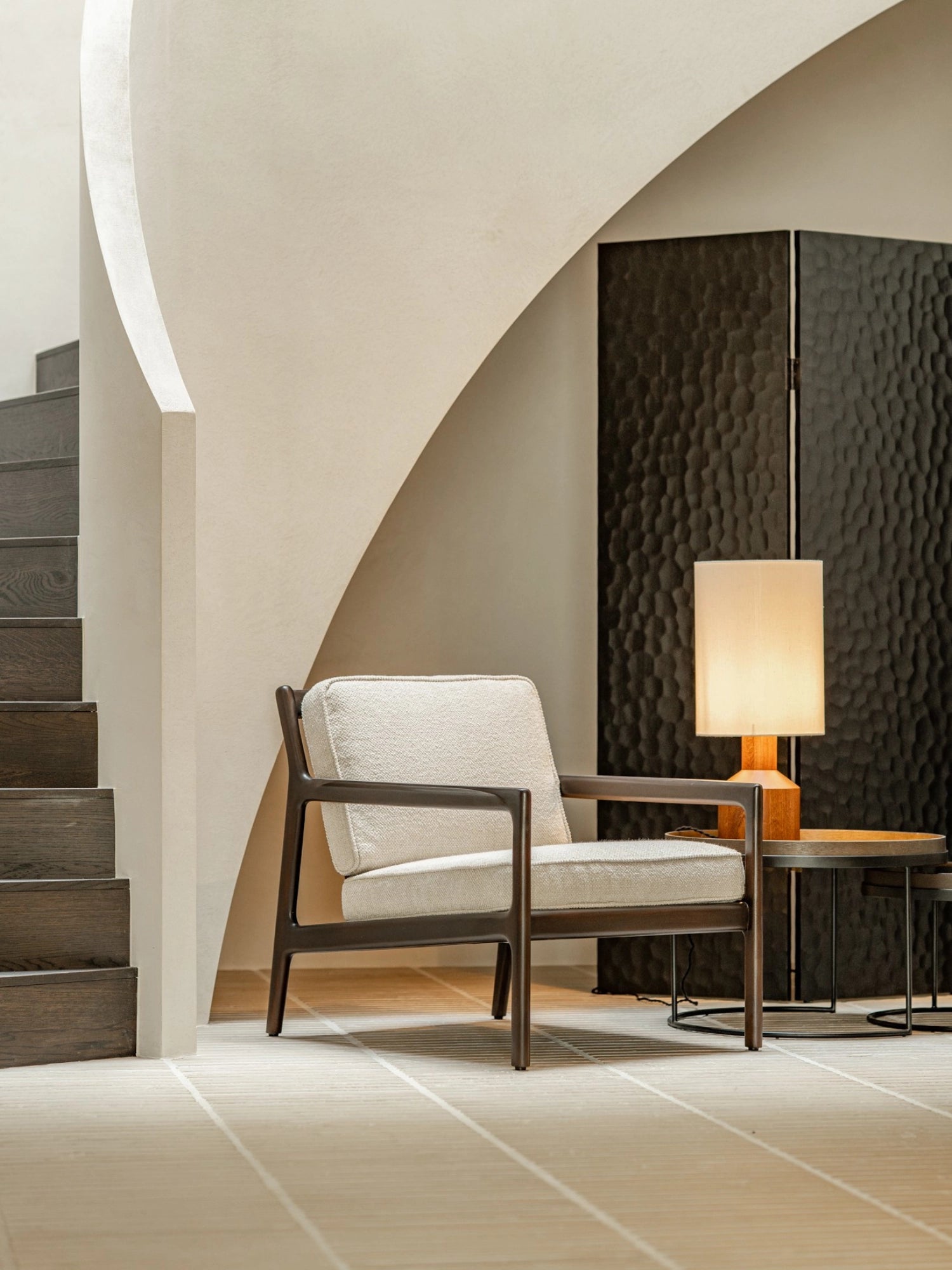 Ethnicraft Chopped Room Divider, a sophisticated mahogany black screen with a distinctive textured pattern, positioned elegantly in a modern living area with a cream armchair and spiral staircase.