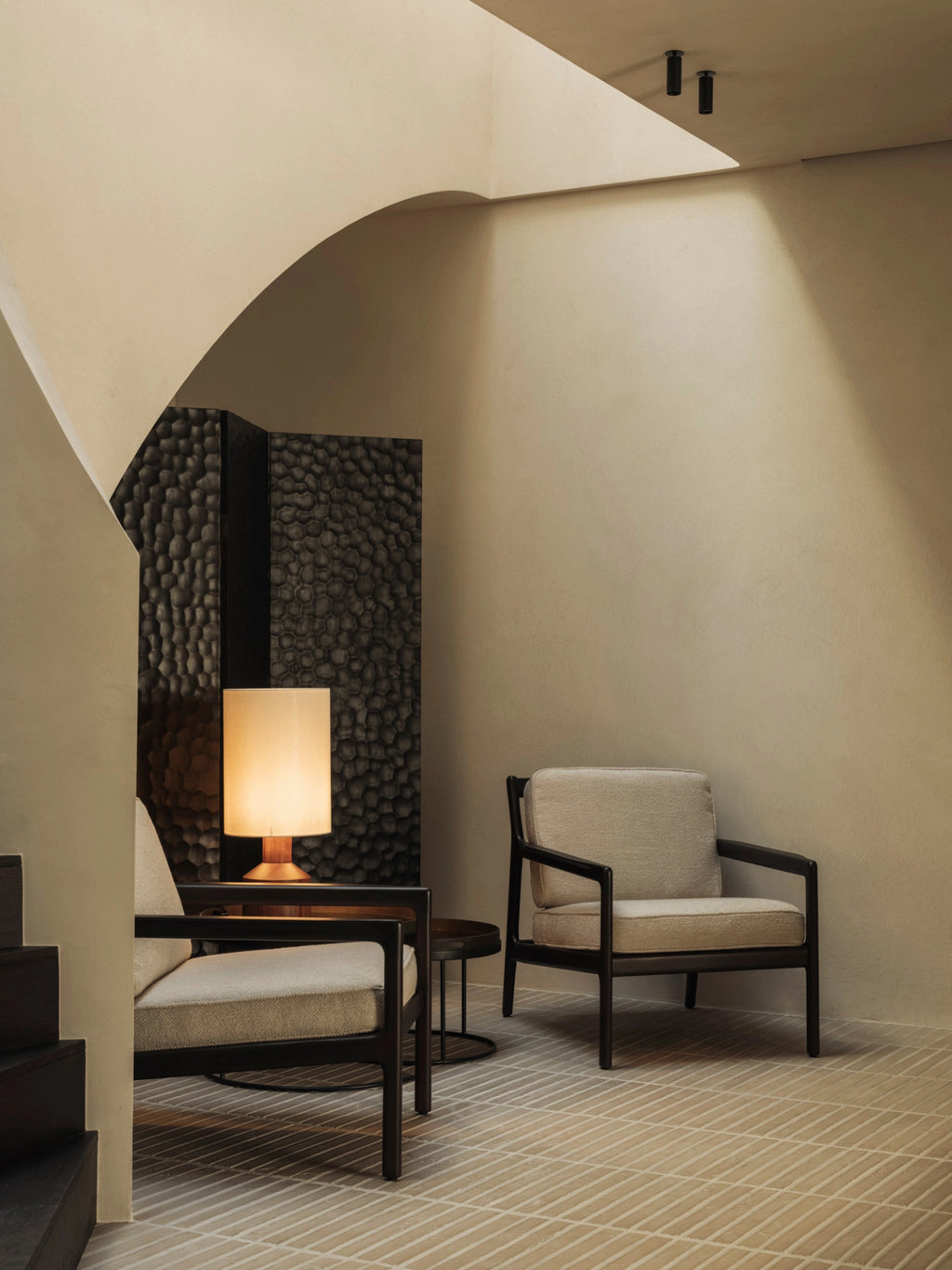 Ethnicraft Chopped Room Divider, a dark mahogany black screen with an intricate, textured pattern, creating a chic backdrop in a contemporary space with two cream armchairs and warm lighting.