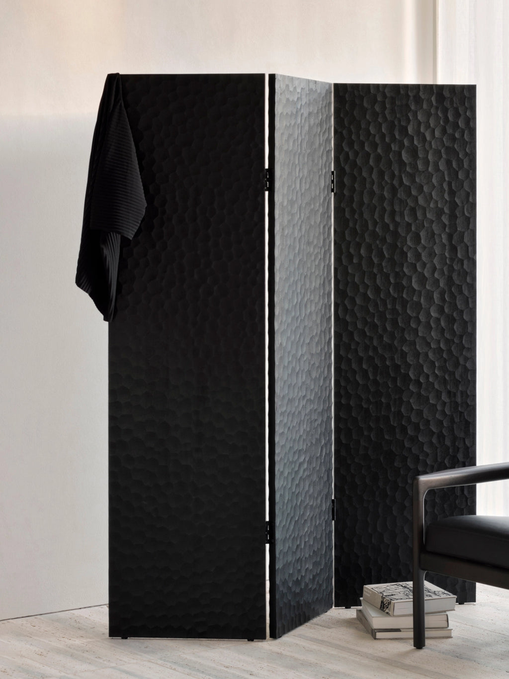 Ethnicraft Chopped Room Divider, a three-panel mahogany black screen with a unique, deeply textured surface, providing a stylish room separation, draped with a black garment in a bright room.