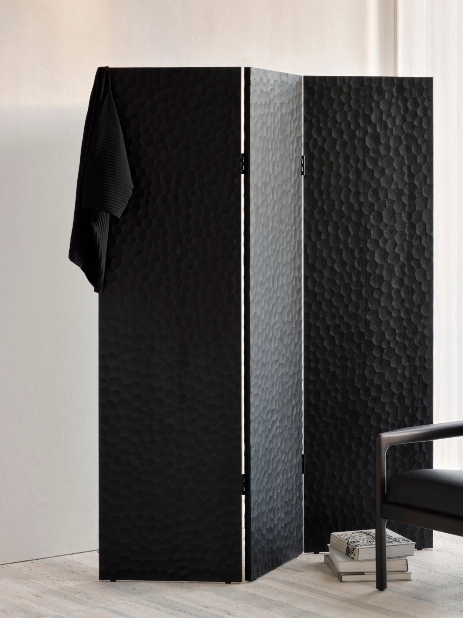Ethnicraft Chopped Room Divider, a three-panel mahogany black screen with a unique, deeply textured surface, providing a stylish room separation, draped with a black garment in a bright room.