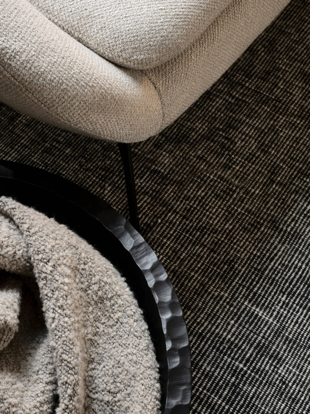 Ethnicraft Chopped XL Bowl, a close-up, overhead view of the large, dark, textured wooden bowl holding a soft, light-colored blanket, placed on a patterned rug next to a cream-colored sofa, emphasizing its tactile quality.