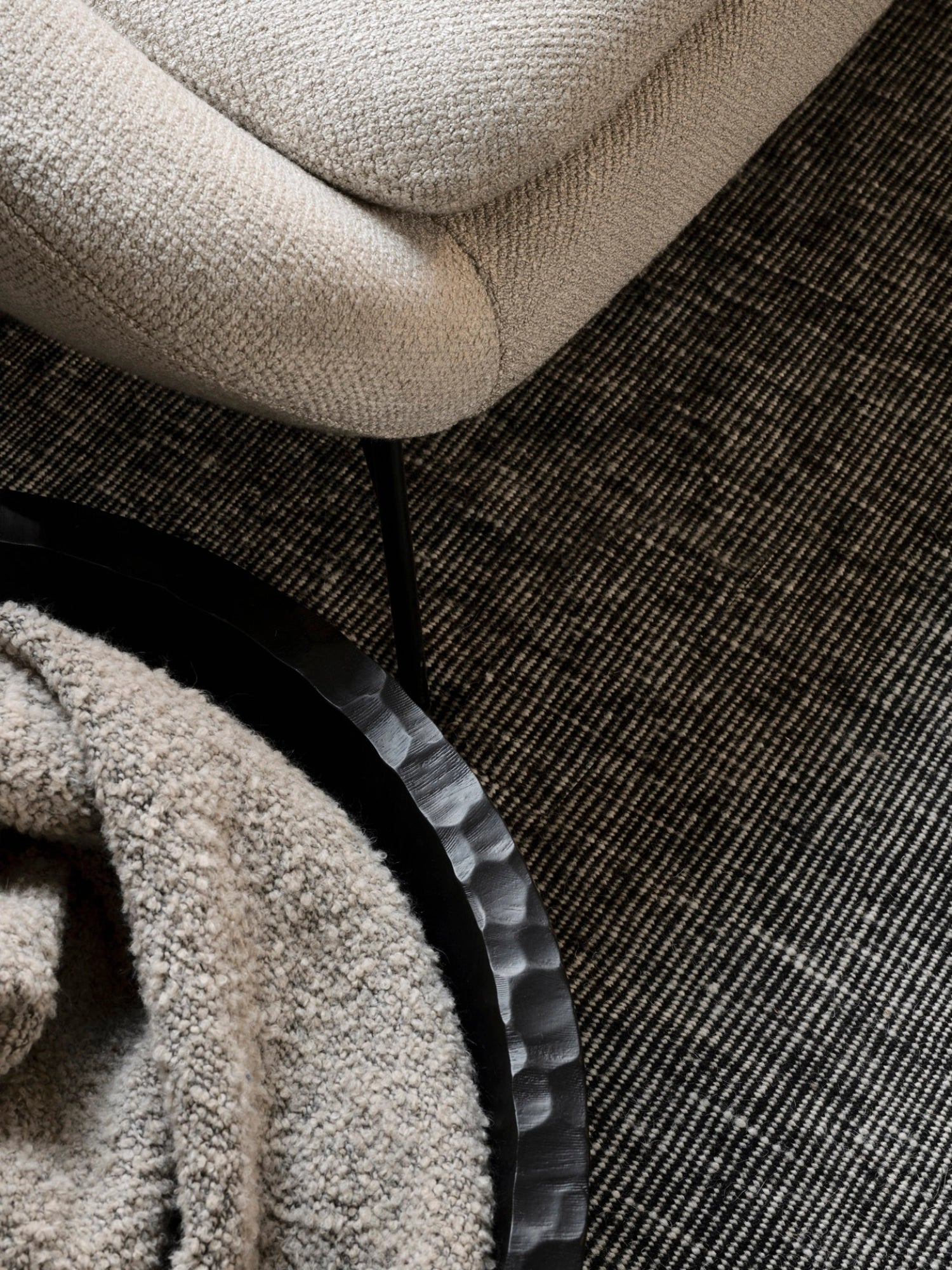 Ethnicraft Chopped XL Bowl, a close-up, overhead view of the large, dark, textured wooden bowl holding a soft, light-colored blanket, placed on a patterned rug next to a cream-colored sofa, emphasizing its tactile quality.