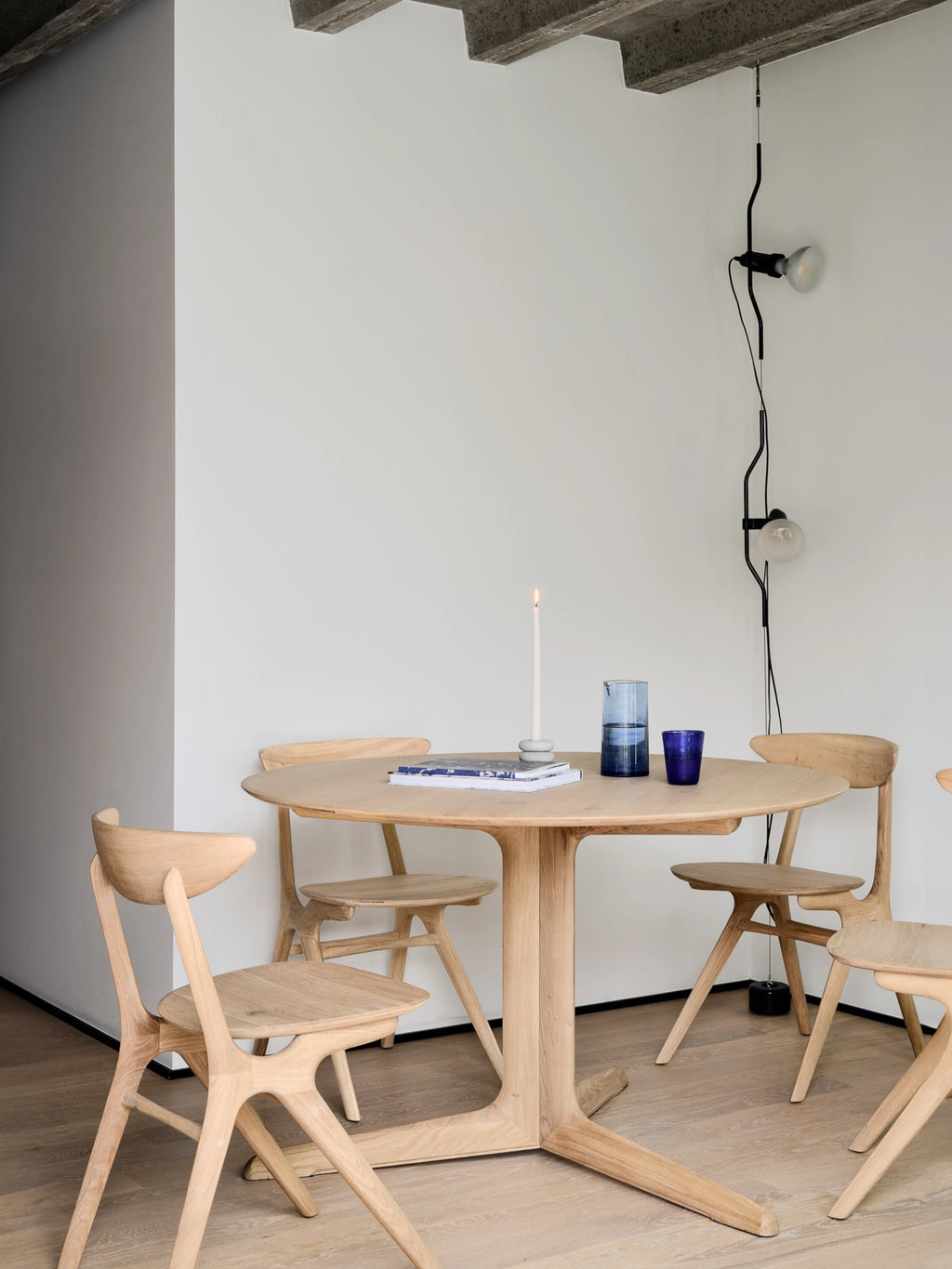 Ethnicraft Corto Dining Table, round oak, presented in a minimalist modern dining room with light-colored walls and concrete ceiling, surrounded by matching wooden chairs.