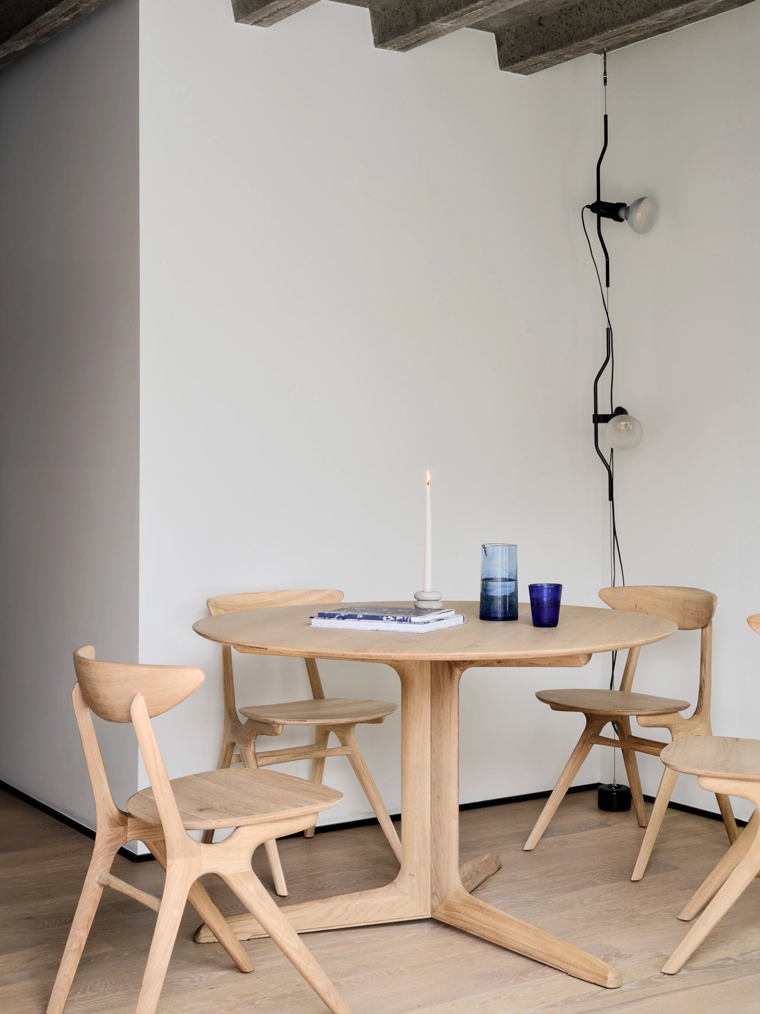 Ethnicraft Corto Dining Table, round oak, presented in a minimalist modern dining room with light-colored walls and concrete ceiling, surrounded by matching wooden chairs.