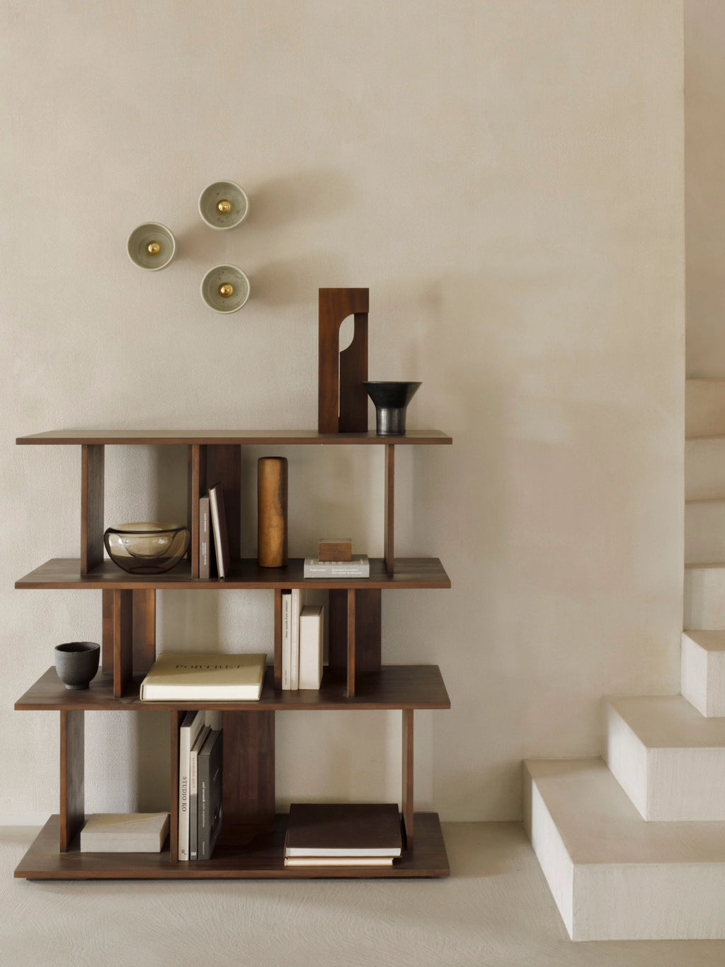 Ethnicraft Irregular Rack, a stylish lifestyle shot featuring the dark brown solid teak wood shelving unit filled with books and decorative objects, positioned against a warm, neutral wall next to a modern staircase, exuding a sophisticated, contemporary aesthetic.