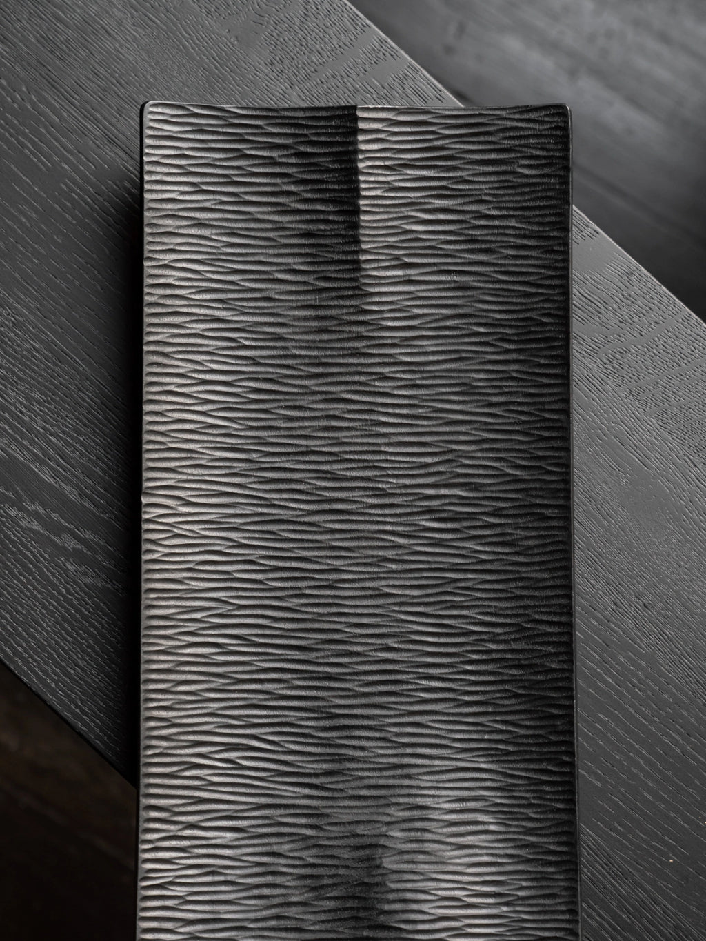 Ethnicraft Japan Board, a top-down lifestyle shot of the rectangular black serving board, resting on a dark wooden surface. The board's intricate wave-like texture is prominently displayed, offering a sophisticated and modern aesthetic for serving.