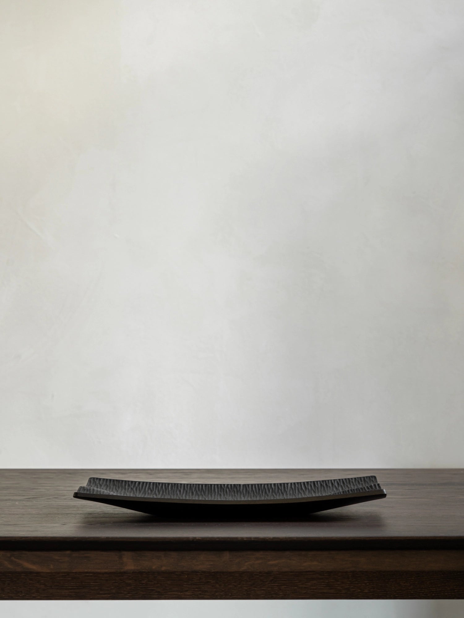 Ethnicraft Japan Board, a minimalist black rectangular serving board with a textured surface, displayed centrally on a rich dark wooden table against a light, textured wall, highlighting its refined aesthetic.