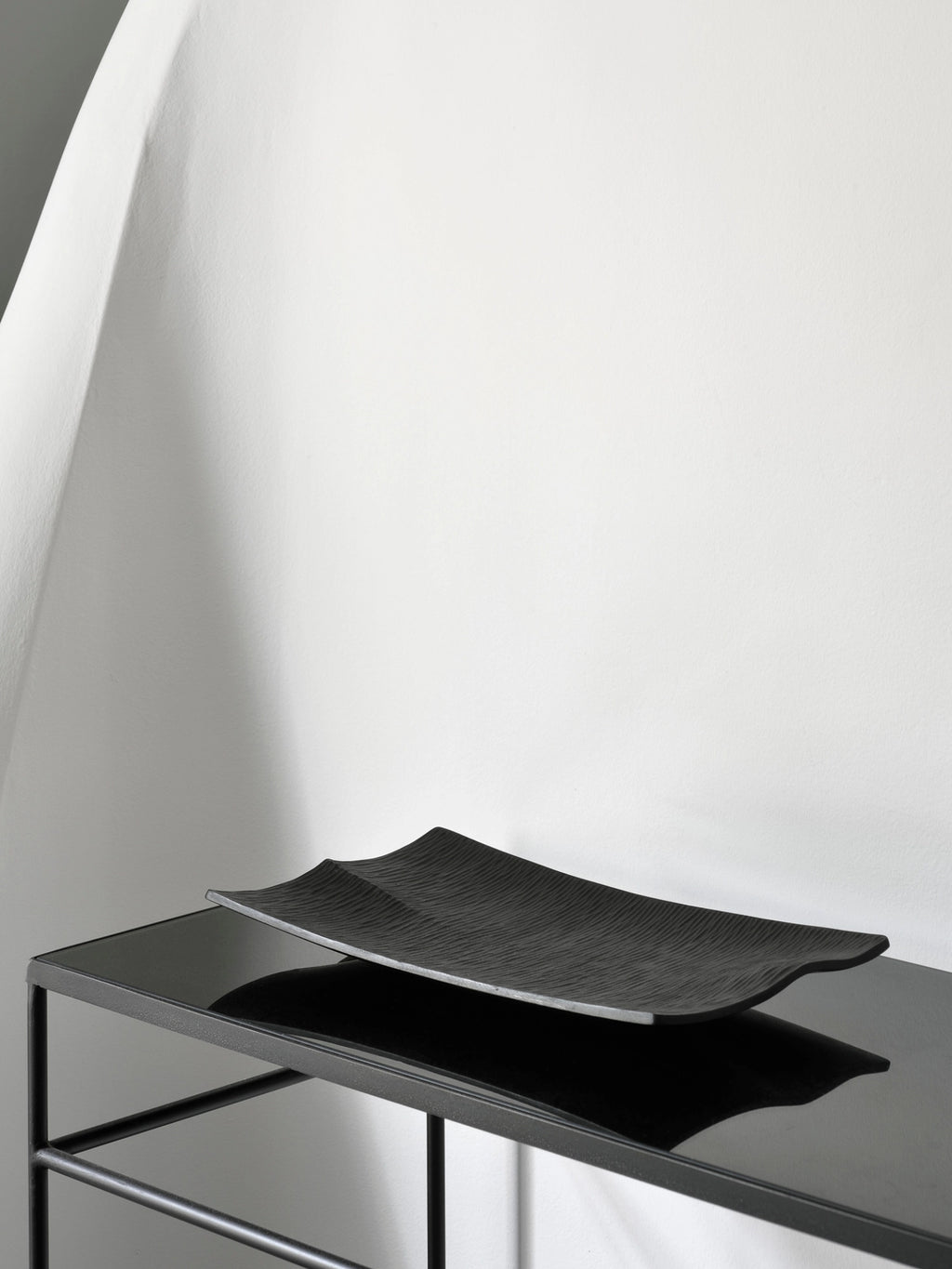 Ethnicraft Japan Board, a sleek black rectangular serving board with a subtle wavy texture, resting on a modern black metal console table against a white wall, showcasing its elegant design.