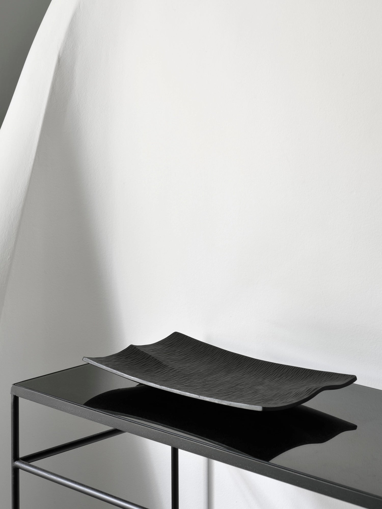 Ethnicraft Japan Board, a sleek black rectangular serving board with a subtle wavy texture, resting on a modern black metal console table against a white wall, showcasing its elegant design.