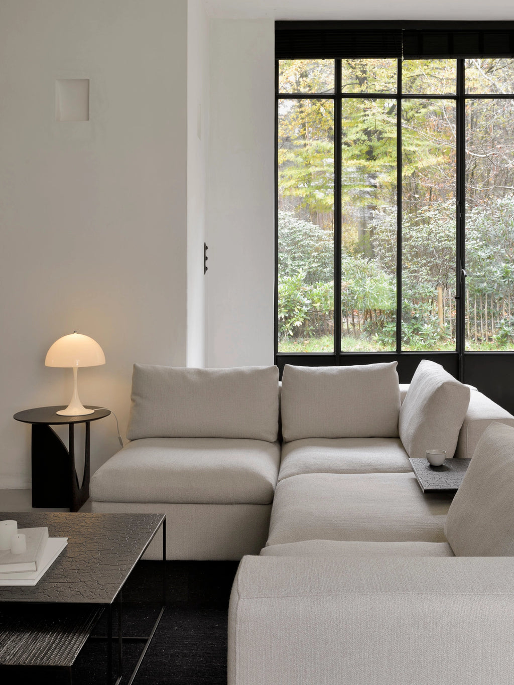 Ethnicraft Mellow Modular Sofa, a stylish light beige L-shaped sectional, featuring clean lines and plush, textured fabric. The modern design is perfectly suited for a contemporary living room with large black-framed windows overlooking lush greenery.