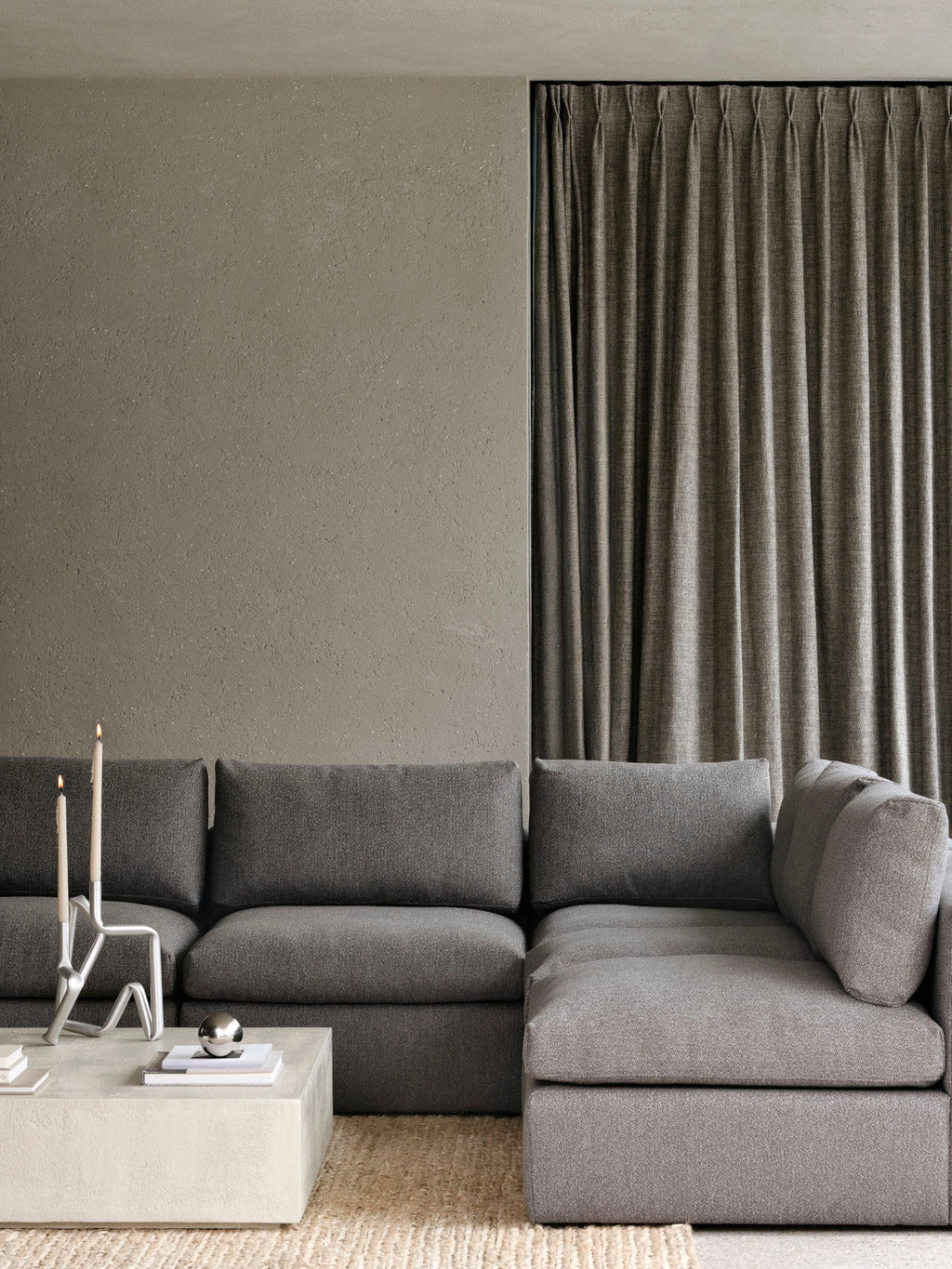 Ethnicraft Mellow Modular Sofa, 1-Seater. Lifestyle shot of the dark grey modular sofa in a modern living room, paired with a concrete coffee table and elegant candles, against a textured wall.