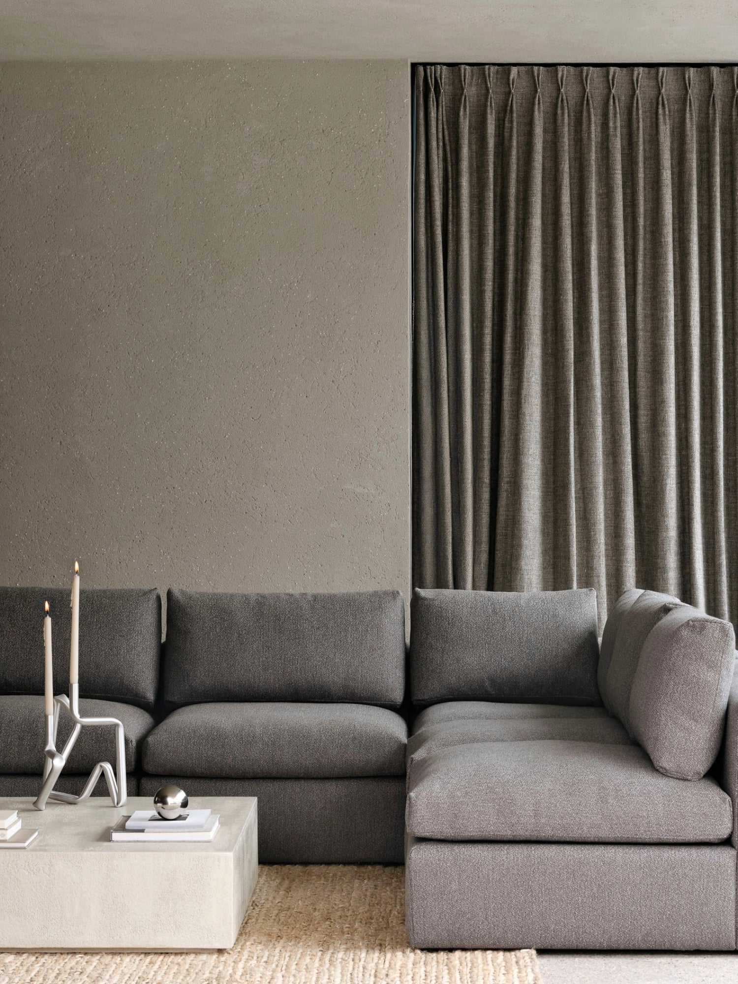 Ethnicraft Mellow Modular Sofa, 1-Seater. Lifestyle shot of the dark grey modular sofa in a modern living room, paired with a concrete coffee table and elegant candles, against a textured wall.
