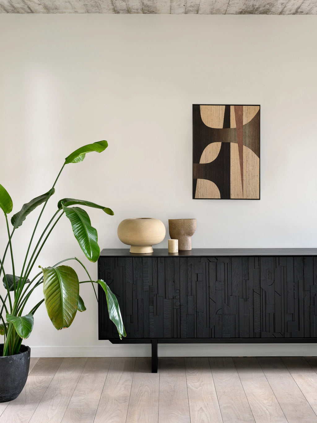 Ethnicraft Mosaic Sideboard in Teak Off-Black, a modern dark wood cabinet with distinctive carved patterns, enhancing a bright living area with a large green plant and abstract art.